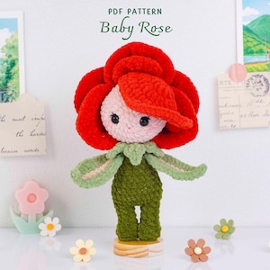 Könnte beinhalten: Gehäkelte Puppe in Form einer Rose, mit rotem Blütenkopf, hellrosa Gesicht und grünen Blättern und Stiel. Die Puppe steht auf einem Holzsockel. Der Text "PDF PATTERN Baby Rose" steht oben.