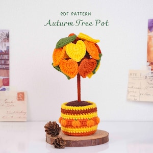 Op de afbeelding: Een haak-PDF-patroon getiteld Auturm Tree Pot met een kleine boom met hartvormige bladeren in geel, oranje en groen. De boom staat in een geel en oranje gestreepte pot met kleine pompoenmotieven, rustend op een ronde houten basis.