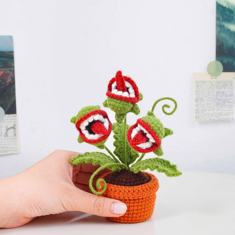 May include: A crocheted plant with three open-mouthed flower heads, each with red, white, and green details. The plant sits in an orange pot with green leaves and stems. The plant is a whimsical, handmade item.