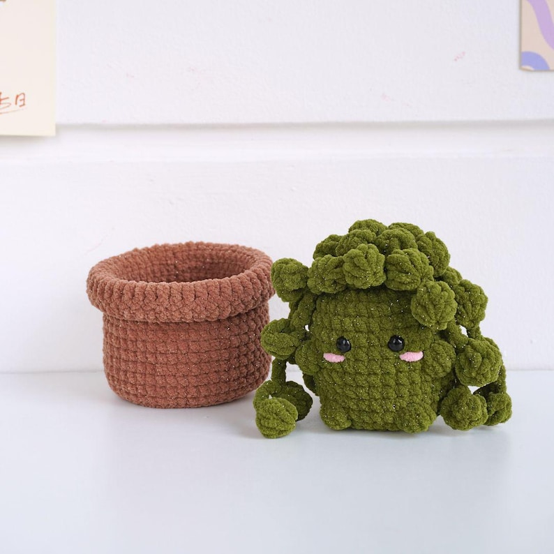 May include: A green crocheted succulent plant with a cute face, sitting in a brown crocheted pot.