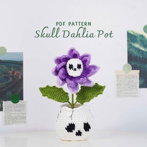 Könnte beinhalten: Eine gehäkelte lila Blume mit einem Totenkopfgesicht in einem weißen Topf mit schwarzen Totenkopfakzenten. Die Blume hat grüne Blätter und ist mit "Skull Dahlia Pot" mit "PDF Pattern" darüber beschriftet.