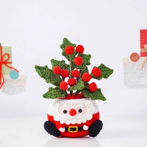 May include: A handmade crochet Santa Claus planter, featuring a red and white outfit with a black belt and boots. The planter holds a green crochet plant with red berries, creating a festive holiday decoration.