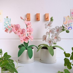 May include: Two crocheted orchid plants in white and pink pots. The orchids have green leaves and are in white and pink flower clusters.