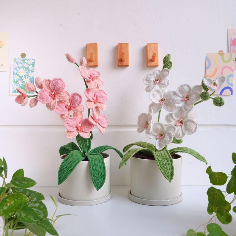 May include: Two crocheted orchid plants in white and pink pots. The orchids have green leaves and are in white and pink flower clusters.