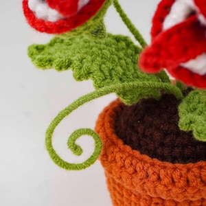 May include: A crocheted plant with red and white flowers, green leaves, and a curly green vine. The plant is in an orange pot with a brown interior. The handmade item is likely a decorative piece.