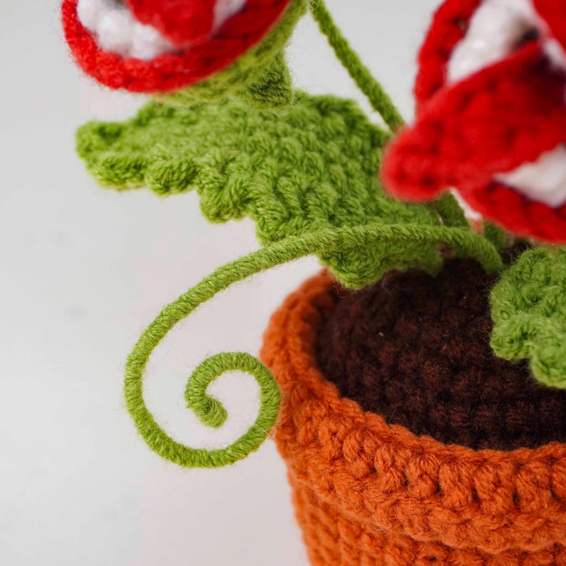 May include: A crocheted plant with red and white flowers, green leaves, and a curly green vine. The plant is in an orange pot with a brown interior. The handmade item is likely a decorative piece.