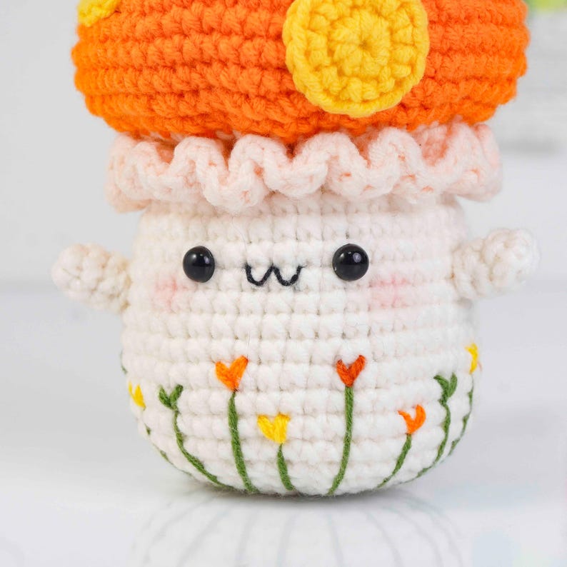 May include: A crocheted mushroom figure with an orange cap and yellow accents. The mushroom has a white body with a smiling face, small arms, and embroidered flowers around the base. The figure is handmade.