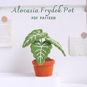 Puede incluir: Una planta Alocasia Frydek de crochet en una maceta de terracota. La planta tiene hojas verdes con venas blancas y está sentada sobre una superficie blanca. El texto "Alocasia Frydek Pot PDF PATTERN" está escrito sobre la planta.