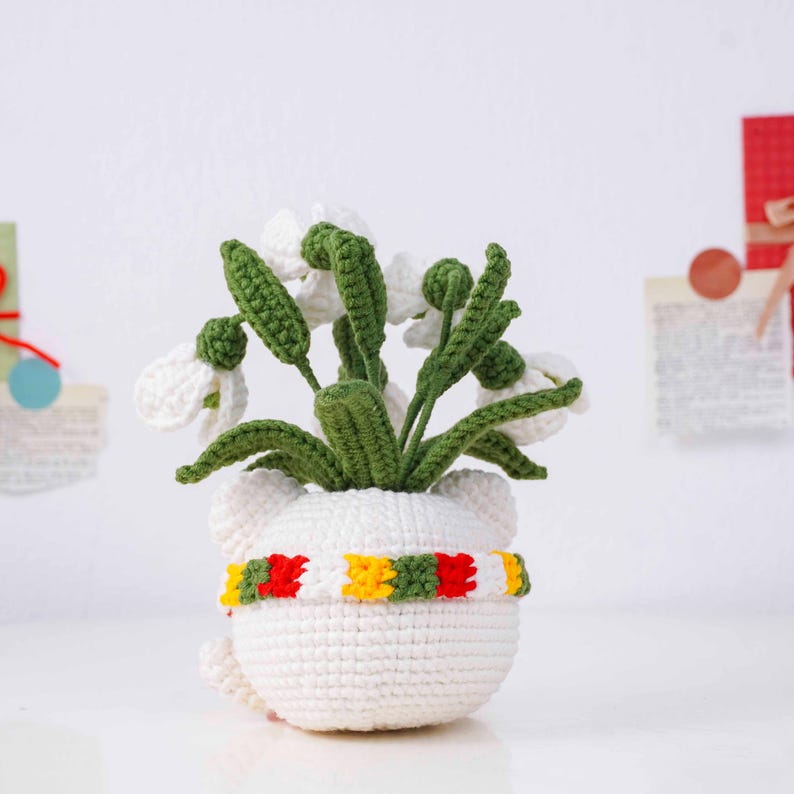 May include: A handmade crochet snowdrop flower arrangement in a white pot with a colorful scarf. The flowers are white with green stems and leaves. The pot has a bear-like shape. The scarf has red, yellow, and green stripes.