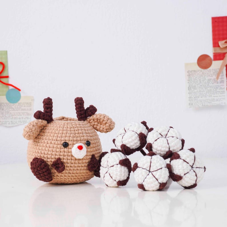 May include: A crocheted reindeer with brown antlers, ears, and feet, and a red nose, sits next to several white and brown crocheted cotton ball ornaments. The reindeer is tan and has black eyes. The items are on a white surface.