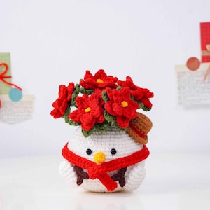 May include: A crocheted snowman with a red scarf and a small brown hat. The snowman's head is topped with a bouquet of red flowers with yellow centers. The snowman has black eyes and a yellow beak.