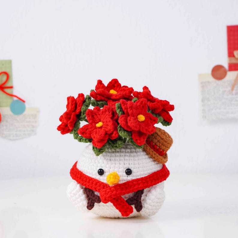 May include: A crocheted snowman with a red scarf and a small brown hat. The snowman's head is topped with a bouquet of red flowers with yellow centers. The snowman has black eyes and a yellow beak.