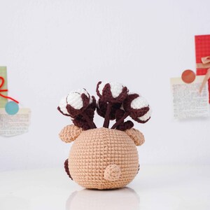 May include: A crocheted reindeer-shaped vase in tan holds several cotton flower stems with white and brown accents. The handmade vase has small ears and a tail. The cotton flowers add a touch of nature to the decorative piece.