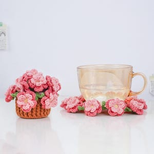 Puede incluir: Una taza de t&eacute; de vidrio transparente con asa se encuentra junto a una peque&ntilde;a cesta llena de flores rosas de ganchillo. Varias flores rosas individuales de ganchillo con hojas verdes est&aacute;n esparcidas alrededor de la taza y la cesta. La cesta es marr&oacute;n.