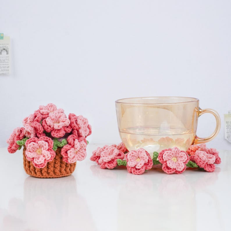 Puede incluir: Una taza de t&eacute; de vidrio transparente con asa se encuentra junto a una peque&ntilde;a cesta llena de flores rosas de ganchillo. Varias flores rosas individuales de ganchillo con hojas verdes est&aacute;n esparcidas alrededor de la taza y la cesta. La cesta es marr&oacute;n.