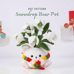 May include: A crocheted white bear pot holding white and green snowdrop flowers. The bear has a colorful scarf and the text "PDF PATTERN Snowdrop Bear Pot" is at the top. Decorative gift boxes are in the background.