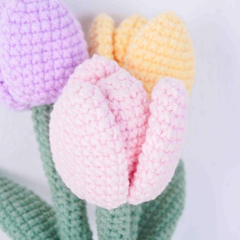 May include: Close-up of three crocheted tulips in shades of pink, yellow, and purple. The flowers have green stems and leaves. The soft texture of the yarn is visible, creating a handmade appearance. The background is plain white.