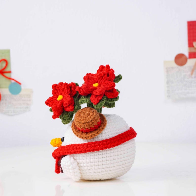 May include: A crocheted snowman figurine with red poinsettia flowers, a brown hat, and a red scarf. The snowman has a yellow beak and is made of white yarn. A festive, handmade decoration.