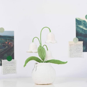 May include: A crocheted lily of the valley plant in a white vase. The plant features three white bell-shaped flowers with green stems and leaves. The vase is also white and has a textured appearance.