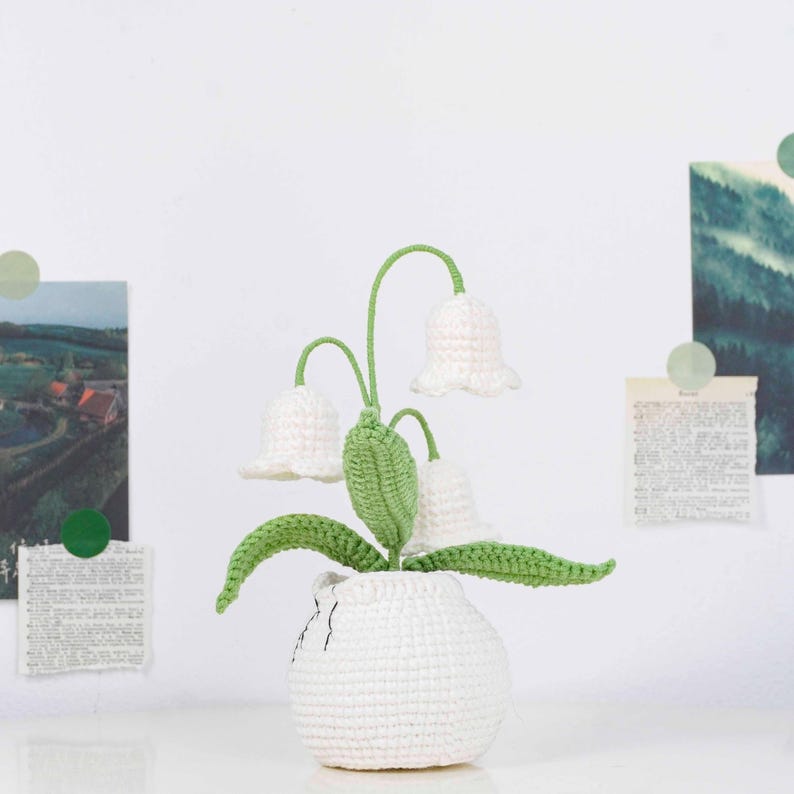 May include: A crocheted lily of the valley plant in a white vase. The plant features three white bell-shaped flowers with green stems and leaves. The vase is also white and has a textured appearance.