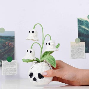 May include: A crocheted white flower arrangement with three ghost-shaped blooms, green stems, and leaves, set in a white skull-shaped pot with black accents. The handmade decor is displayed on a white surface.
