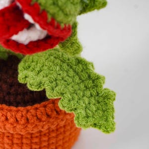 May include: A crocheted plant with a large red and white mouth, green leaves, and an orange pot. The plant is made of yarn and has a textured appearance. The leaves have a scalloped edge.