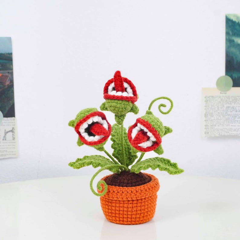 May include: A crocheted plant with three open-mouthed flower heads, each with a red tongue and white teeth. The green leaves and stems sit in an orange pot. The plant is a whimsical, handmade creation.