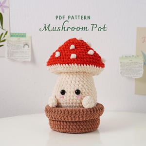 May include: A handmade crochet mushroom pot with a red cap dotted with white spots. The mushroom has a cream-colored stem and is nestled in a brown pot. The text "PDF PATTERN Mushroom Pot" is above the mushroom.