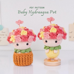 May include: Two crocheted figures with pink and yellow hydrangea flower heads, small black eyes, and rosy cheeks. One figure sits in a brown textured pot, while the other stands on a round wooden base. Text reads: PDF PATTERN Baby Hydrangea Pot.