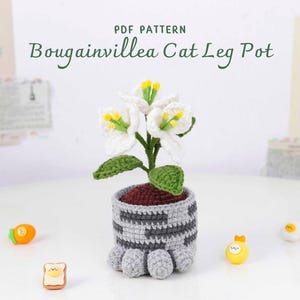 May include: A crocheted Bougainvillea Cat Leg Pot with white flowers, green leaves, and a gray and white patterned pot. The pot has gray cat paw feet. The text "PDF PATTERN Bougainvillea Cat Leg Pot" is at the top.