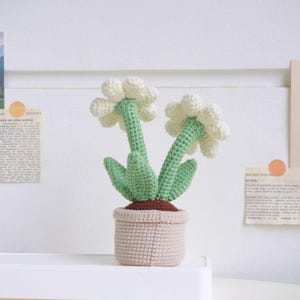 Puede incluir: Una planta de crochet con flores blancas y hojas verdes en una maceta beige. La planta es un art&iacute;culo artesanal hecho a mano.