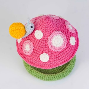 May include: A handmade crochet mushroom hat in bright pink with white polka dots and a green brim. A yellow snail with a blue body is attached to the side of the hat. The hat is designed for babies and children.