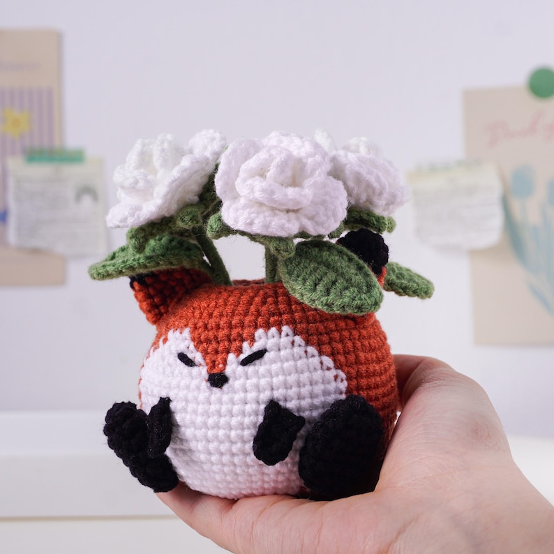 May include: A crocheted fox-shaped planter in orange, white, and black, holding white crocheted roses with green leaves. The fox has closed eyes and black paws. A charming handmade item for home decor.