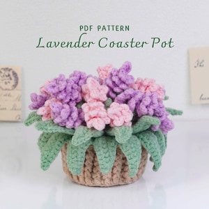 May include: A crocheted lavender coaster pot featuring purple and pink flowers and green leaves. The pot is light brown. The text "PDF PATTERN" and "Lavender Coaster Pot" are displayed above the pot.