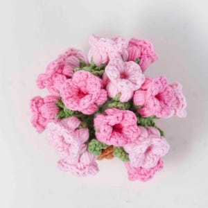 以下が含まれることがあります： ピンク色のグラデーションの、手作りのかぎ針編みの花のブーケ。緑色の葉が付いています。花は円形に配置され、ふっくらとした質感のある外観を作り出しています。背景は無地の白です。