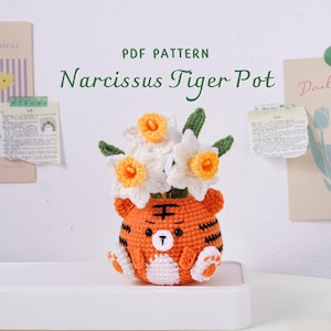 以下が含まれることがあります： 黒い縞模様と可愛らしい顔の、かぎ針編みのオレンジ色のトラのポットには、白と黄色の水仙が入っています。「PDF PATTERN Narcissus Tiger Pot」という文字がポットの上にあります。ポットは白い棚の上にあります。