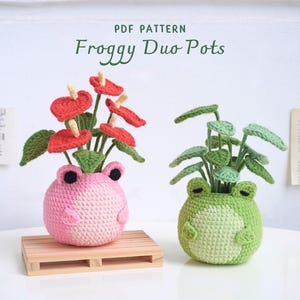 May include: Two crocheted frog-shaped plant pots, one pink with red flowers and one green with green leaves. The pots are on a white surface with the text "PDF PATTERN Froggy Duo Pots" above them.