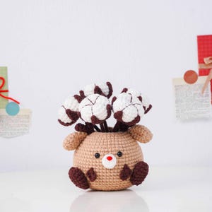 May include: A handmade crochet reindeer vase in tan with brown accents, holding white and brown cotton flower stems. The reindeer has a red nose and black eyes. The vase is sitting on a white surface.
