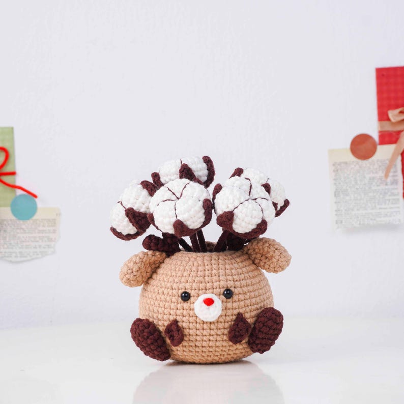 May include: A handmade crochet reindeer vase in tan with brown accents, holding white and brown cotton flower stems. The reindeer has a red nose and black eyes. The vase is sitting on a white surface.