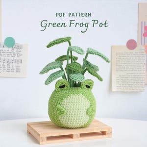 May include: A crochet pattern for a green frog pot with a plant inside. The pattern is called "Green Frog Pot" and is available as a PDF download.