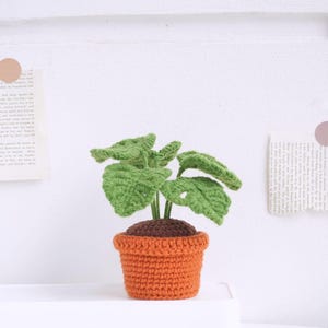 May include: A crocheted plant with green leaves in an orange pot. The plant is a realistic representation of a houseplant.