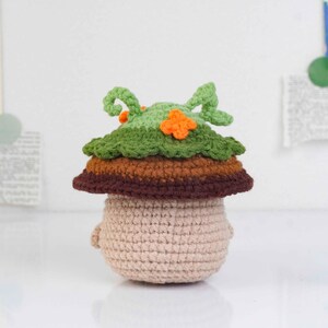 May include: A crocheted mushroom figure with a beige body, a brown and orange midsection, and a green leafy top with orange accents. The handmade amigurumi has small arms and is set against a white background.