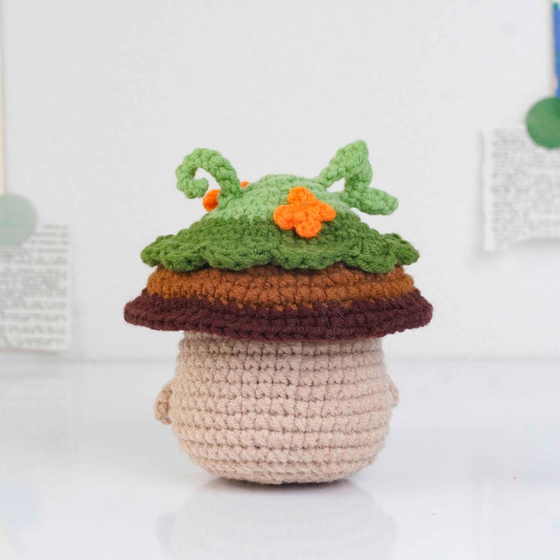 May include: A crocheted mushroom figure with a beige body, a brown and orange midsection, and a green leafy top with orange accents. The handmade amigurumi has small arms and is set against a white background.