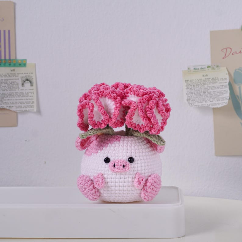 May include: A handmade crochet pig with pink and white spots, featuring a pair of large pink and white flowers on its head. The pig has small black eyes and pink feet. The craft item is sitting on a white surface.