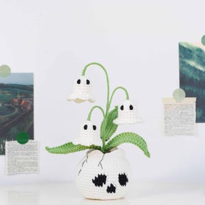 May include: A crocheted decorative plant with three white ghost-shaped flowers with black eyes and green stems and leaves. The plant is in a white crocheted pot with a skull design. The background includes paper clippings and green circular stickers.