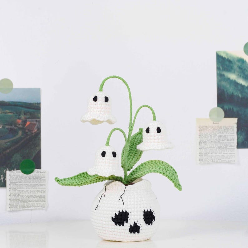 May include: A crocheted decorative plant with three white ghost-shaped flowers with black eyes and green stems and leaves. The plant is in a white crocheted pot with a skull design. The background includes paper clippings and green circular stickers.