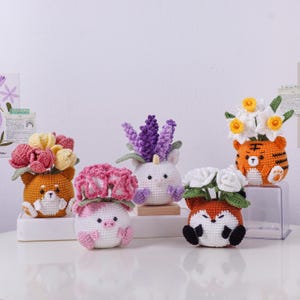May include: A collection of five crocheted animal planters, each holding different colorful flowers. The planters are shaped like a Shiba Inu, pig, unicorn, fox, and tiger. The flowers include pink, yellow, and white blooms, and purple lavender.