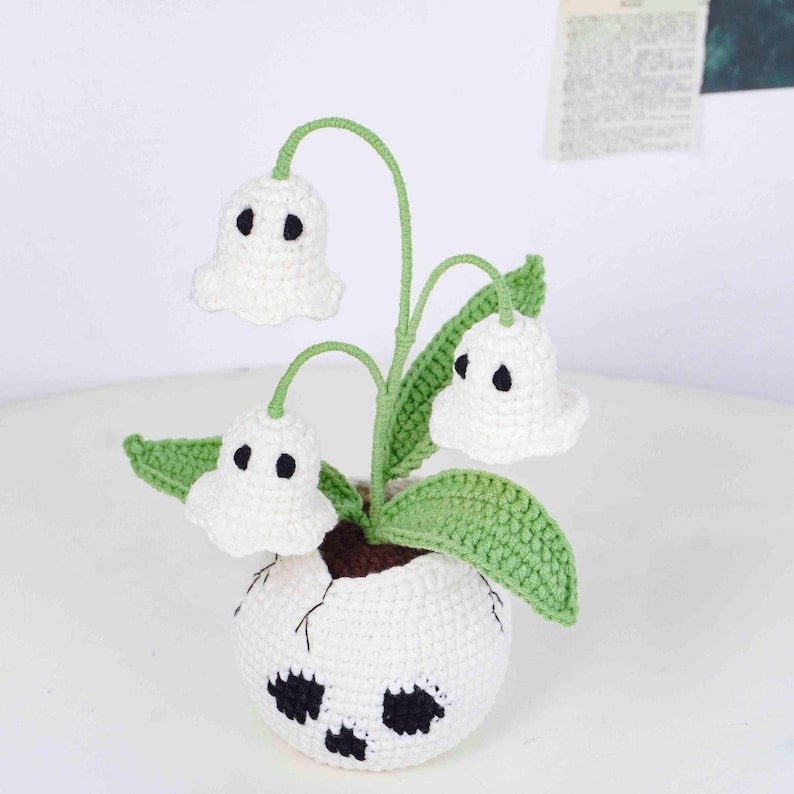 May include: A handmade crochet plant featuring white ghost-shaped flowers with black eyes, green stems and leaves, and a white pot with black accents. The plant is a whimsical, decorative item.