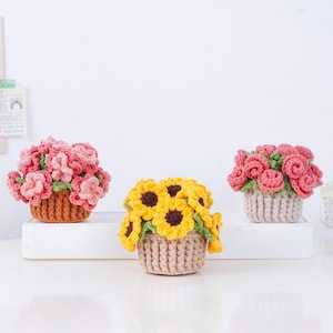 Puede incluir: Tres arreglos florales de ganchillo en cestas peque&ntilde;as. Los arreglos incluyen rosas rosas, girasoles amarillos y flores rosas. Las cestas son en tonos marrones y beige, sobre un fondo blanco.