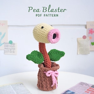 May include: A crocheted Pea Blaster plush toy, a character from the video game Plants vs. Zombies. The toy is yellow with a pink mouth and green leaves. It is sitting in a brown crocheted pot.